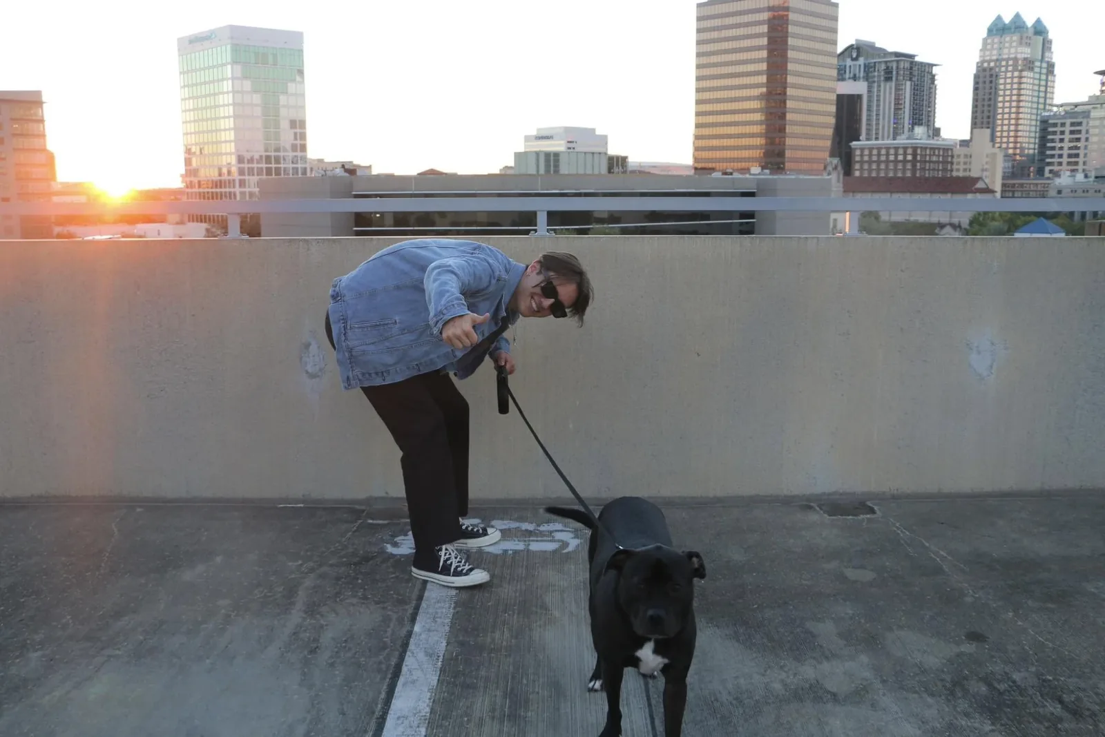 Niko on a rooftop at sunset, smiling and giving a thumbs-up, with his black dog Brier on a leash